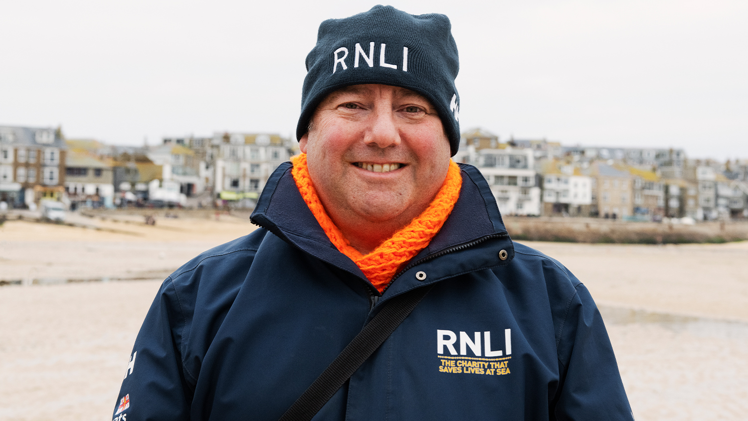 RNLI face-to-face fundraiser David, smiling in a blue RNLI jacket and hat, with Porthmeor Beach, St Ives, in the background