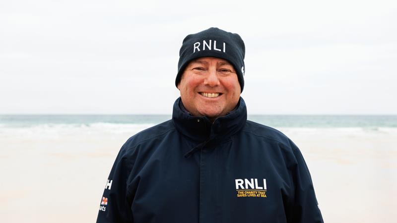 RNLI face-to-face fundraiser David, smiling in a blue RNLI jacket and hat, with Porthmeor Beach, St Ives, in the background