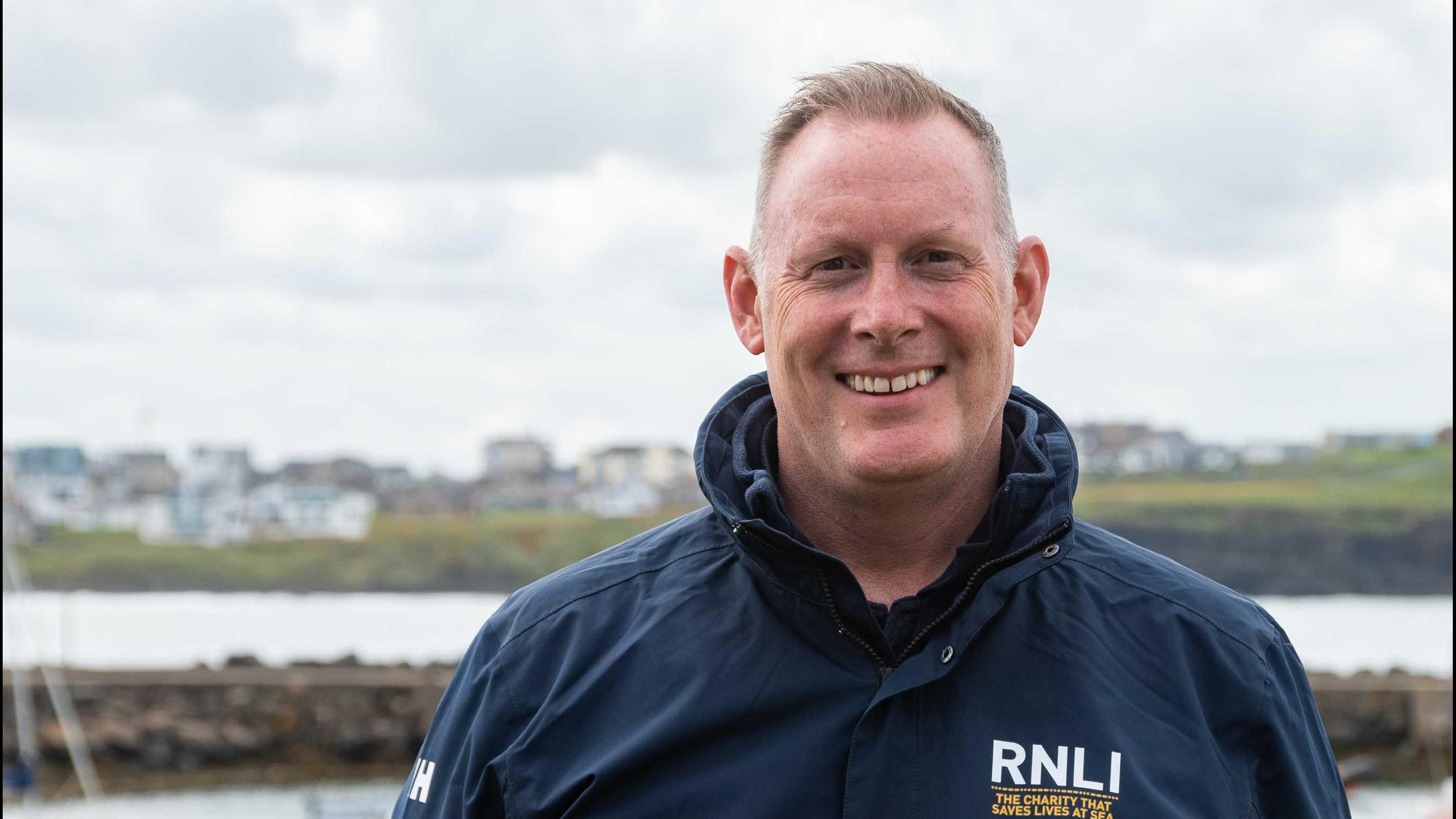 RNLI Face to Face fundraiser Richard Pakenham stood smiling at the camera, wearing a dark blue RNLI jacket at Portrush beach, Northern Ireland.