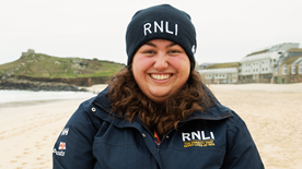 Maya Masi, RNLI face-to-face fundraiser smiling wearing a blue RNLI jacket and beanie hat on the beach
